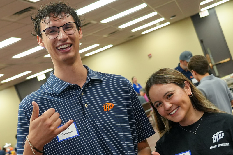 Individual stands at a podium delivering welcome remarks during a UTEP Engineering homecoming event, pointing toward a presentation screen displaying the word “Welcome” beside orange, blue, and silver balloon decorations bearing the College of Engineering logo.